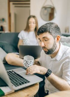 A couple works remotely with laptops in a cozy living room setting, enjoying coffee.