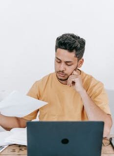 Concentrated young Hispanic male remote specialist in casual wear sitting at table with laptop and examining financial reports while working online in home office